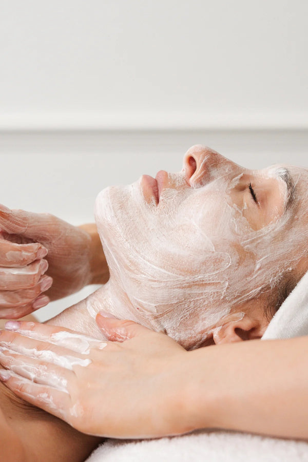 Person receiving a facial treatment with a white mask on a neutral background